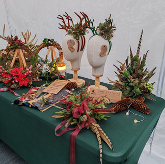 Christmas-themed display with antler headbands on mannequins, wreaths, pinecones, and lights on a green tablecloth. Signs read "Fine Feathers."
