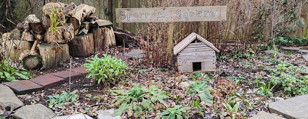 Woodpile and birdhouse in garden labeled "Sparrow Square." Surrounded by rocks, greenery, and a wooden fence. Calm and natural setting.