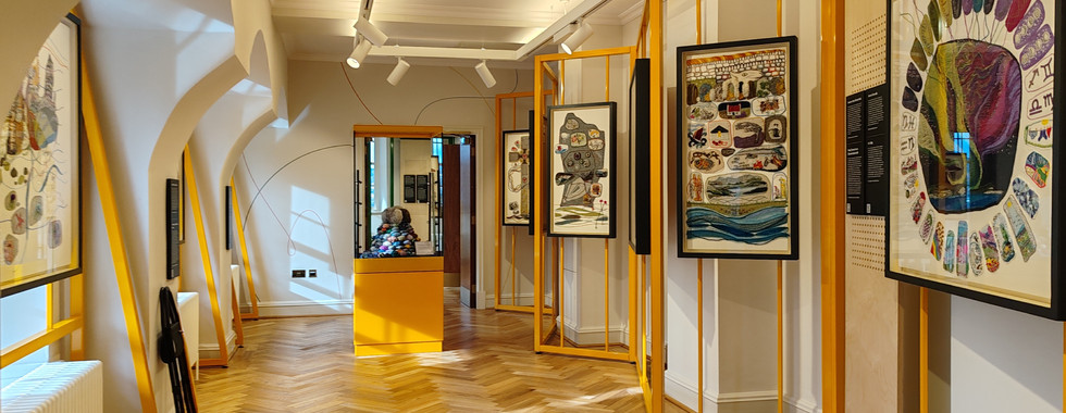 Full room view of tapestry exhibition with colourful tapestries in yellow frames on beige walls. Wooden floor;  bright, well-lit space.