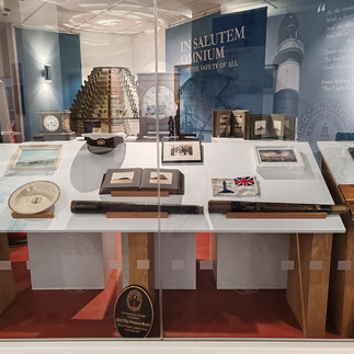 Museum exhibit with nautical items: telescope, captain's hat, maps, and books. Background text reads "IN SALUTEM OMNIUM". Blue and red tones.