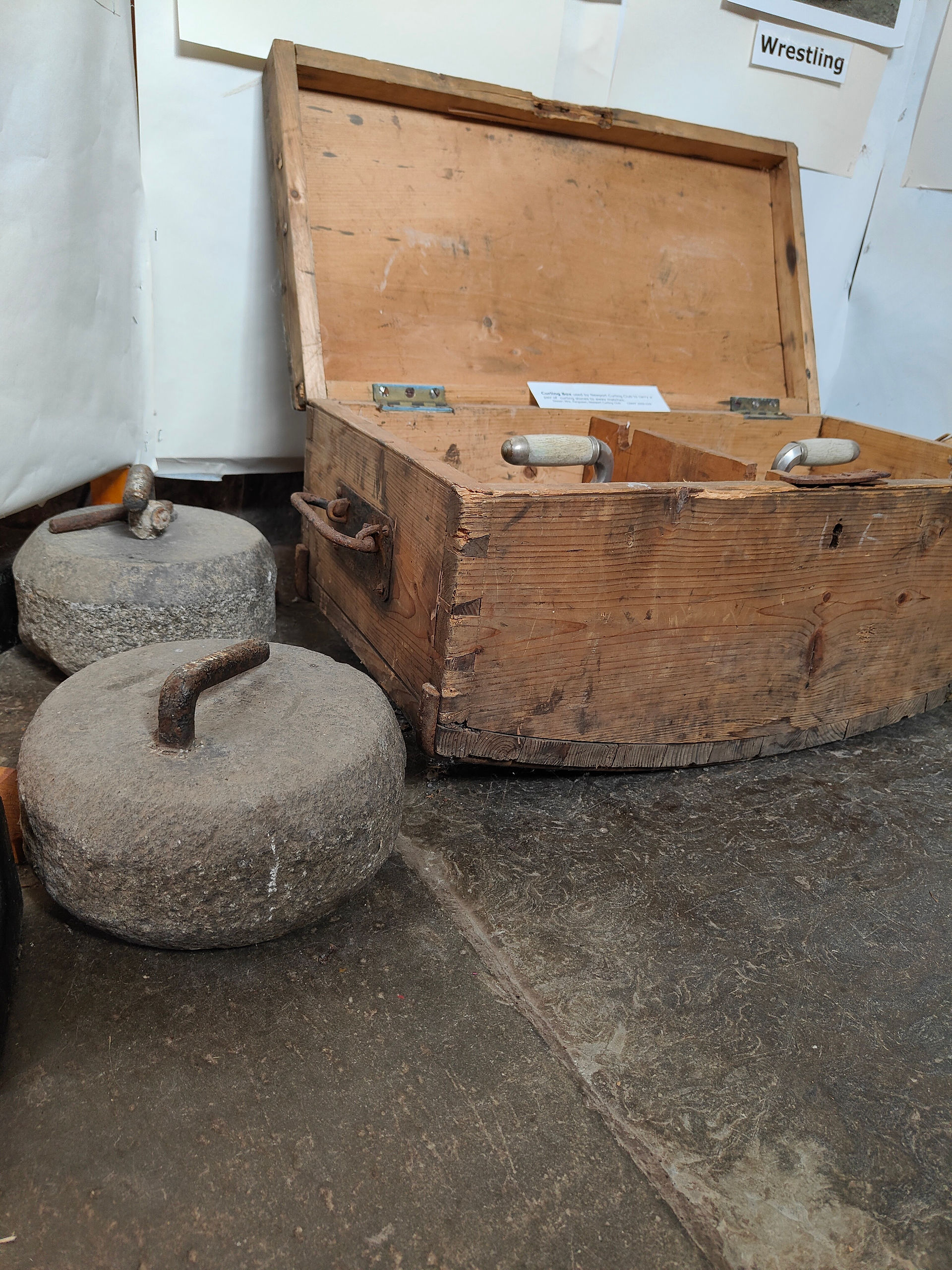 Two antique curling stones on a stone floor next to a wooden chest.