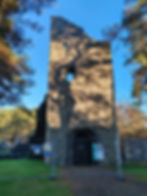 Old stone tower at Cross Kirk with tree shadows and barred entrance. Signs read "Dangerous Masonry" and "Keep Out." Surrounded by trees, under blue sky.