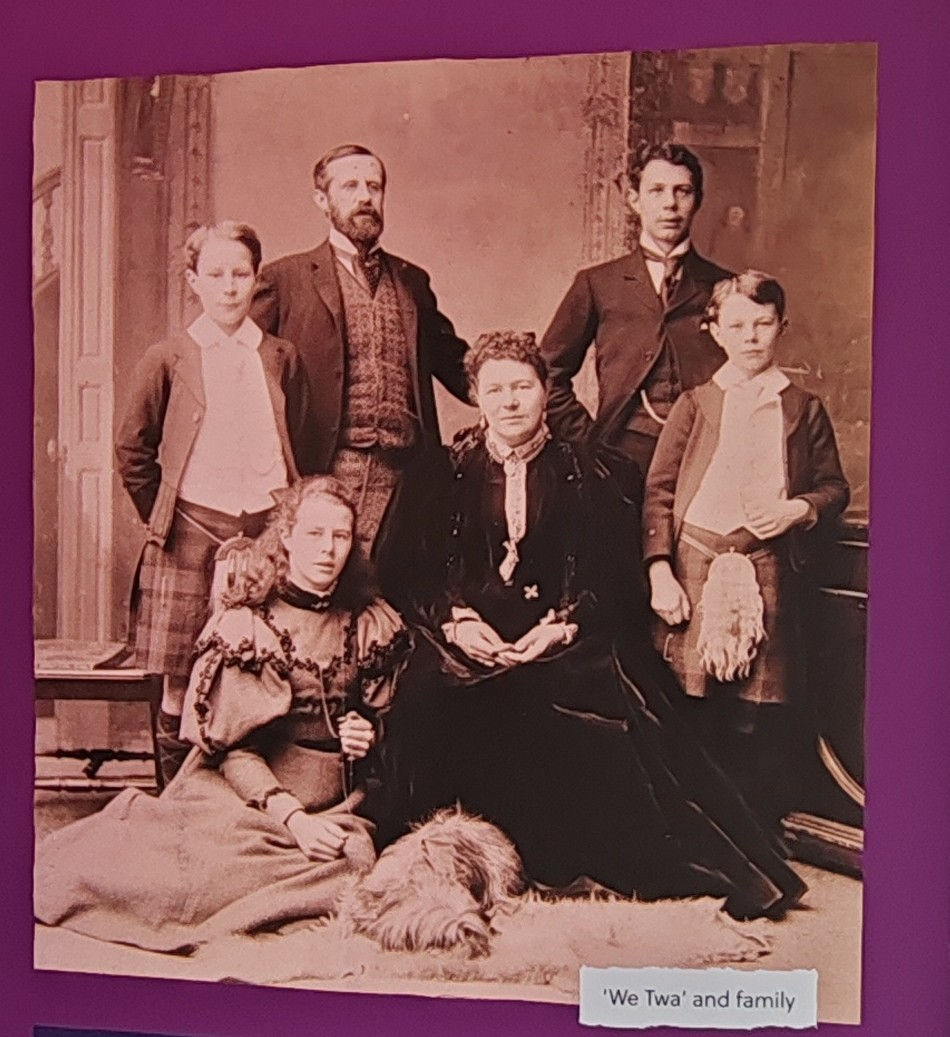 A vintage family portrait of Lord and Lady Aberdeen with their four children in formal attire. A dog lies in front on a rug. Sepia tone with ornate background. Text reads, “We Twa’ and family.”