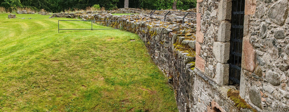View as seen looking out of a window at Huntly Castle of a building with barred window, moss-covered walls, and grassy landscape. Trees in the background under a cloudy sky. Peaceful mood.