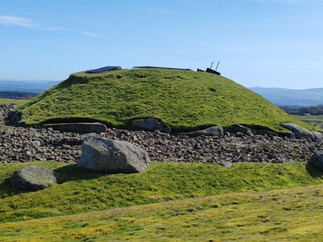 Grassy ancient burial mound with rocks and metal rails on top, under a clear blue sky with distant hills.