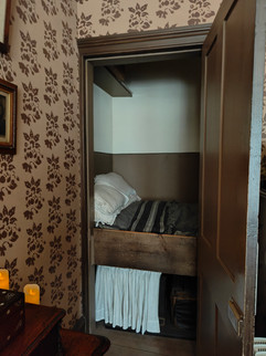 Small wooden bed in a cosy room with floral wallpaper. Bed has white and striped linens. Soft candlelight on a nearby table.