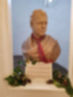 Bust of Walter Scott with a red plaid scarf, placed in a white room. Text plaque below, greenery and pine cones in the foreground.