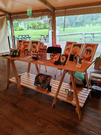 Edinburgh Zoo gift stall under a tent with animal posters, mugs and calendars, overlooking picnic tables and a green park; exit sign above.
