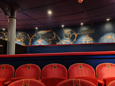 Empty theater interior with red velvet seats and ornate blue walls featuring gold floral patterns. The ceiling is dimly lit.