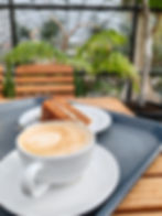 A cappuccino and carrot cake slice on a table in a greenhouse cafe, with wooden chairs and lush green plants in the background.