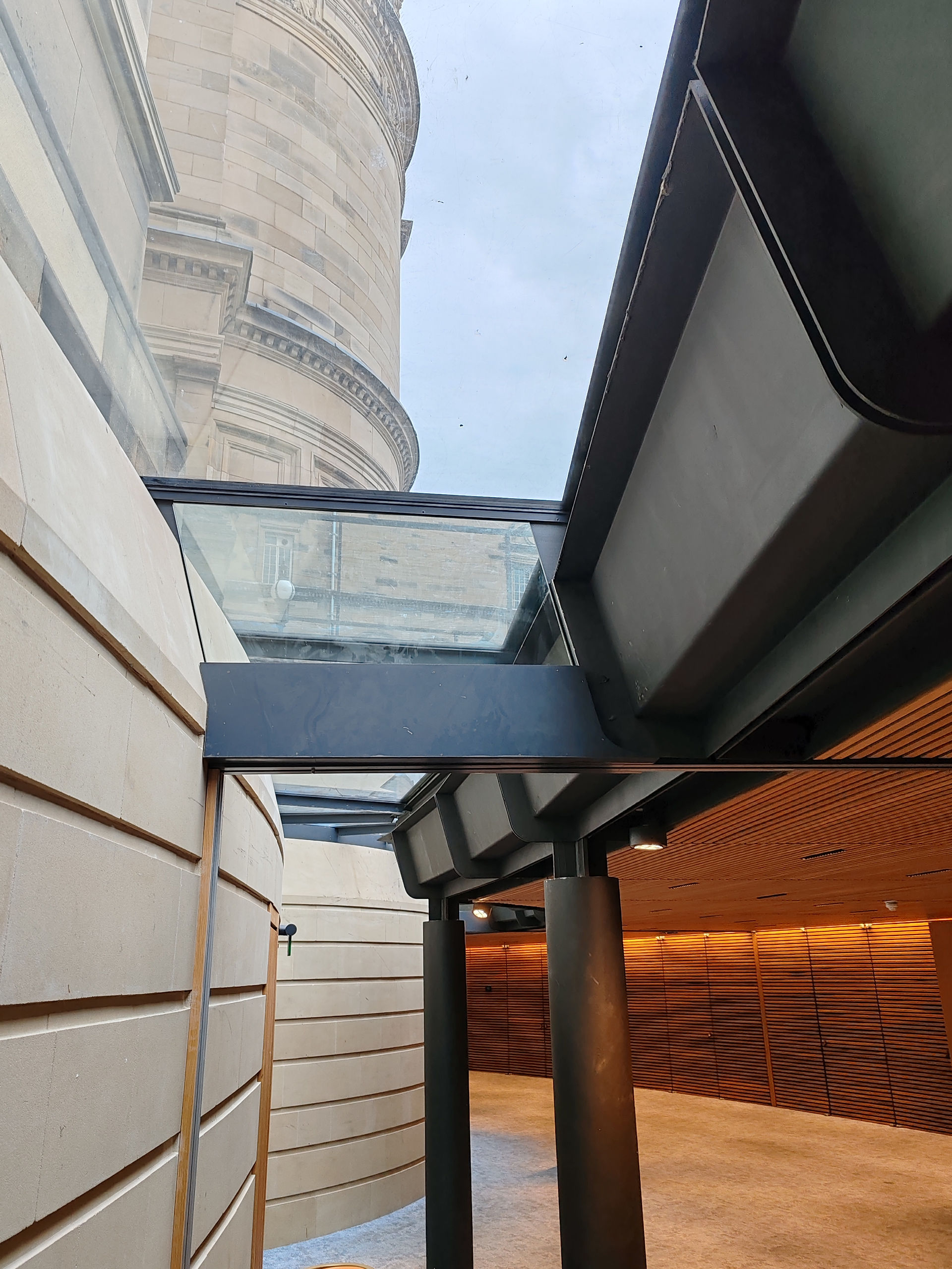 Modern architectural corridor with stone walls and glass ceiling, set against a tall, curved building exterior. No people present.