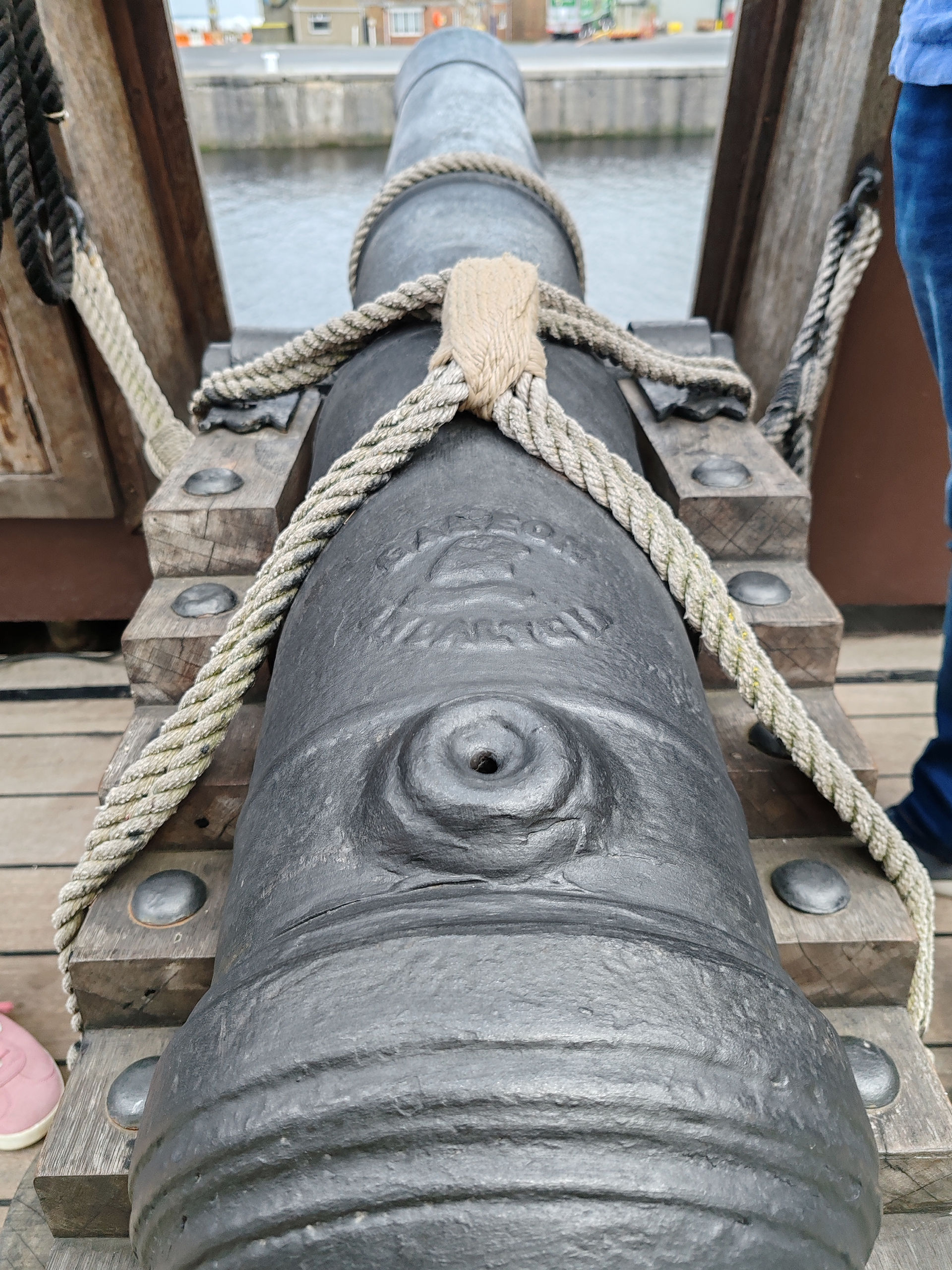 Close-up of a black cannon lashed with rope on a wooden carriage by the water, with a dock and a person in blue at right.