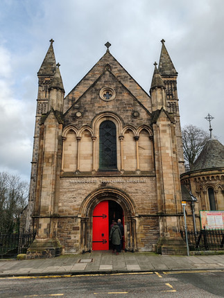 A person enters the Mansfield Traquair Centre, featuring a stone façade with two towers and a bright red door. Overcast sky above.