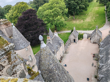 Aerial view of a stone courtyard surrounded by castle towers with conical roofs. Lush green trees and a grassy path in the background.