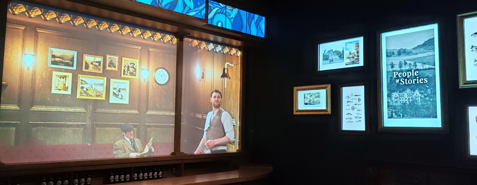 Dimly lit room with framed photos, depicting two men, one with a saxophone. Screen displays text: "People & Stories." Cosy atmosphere.