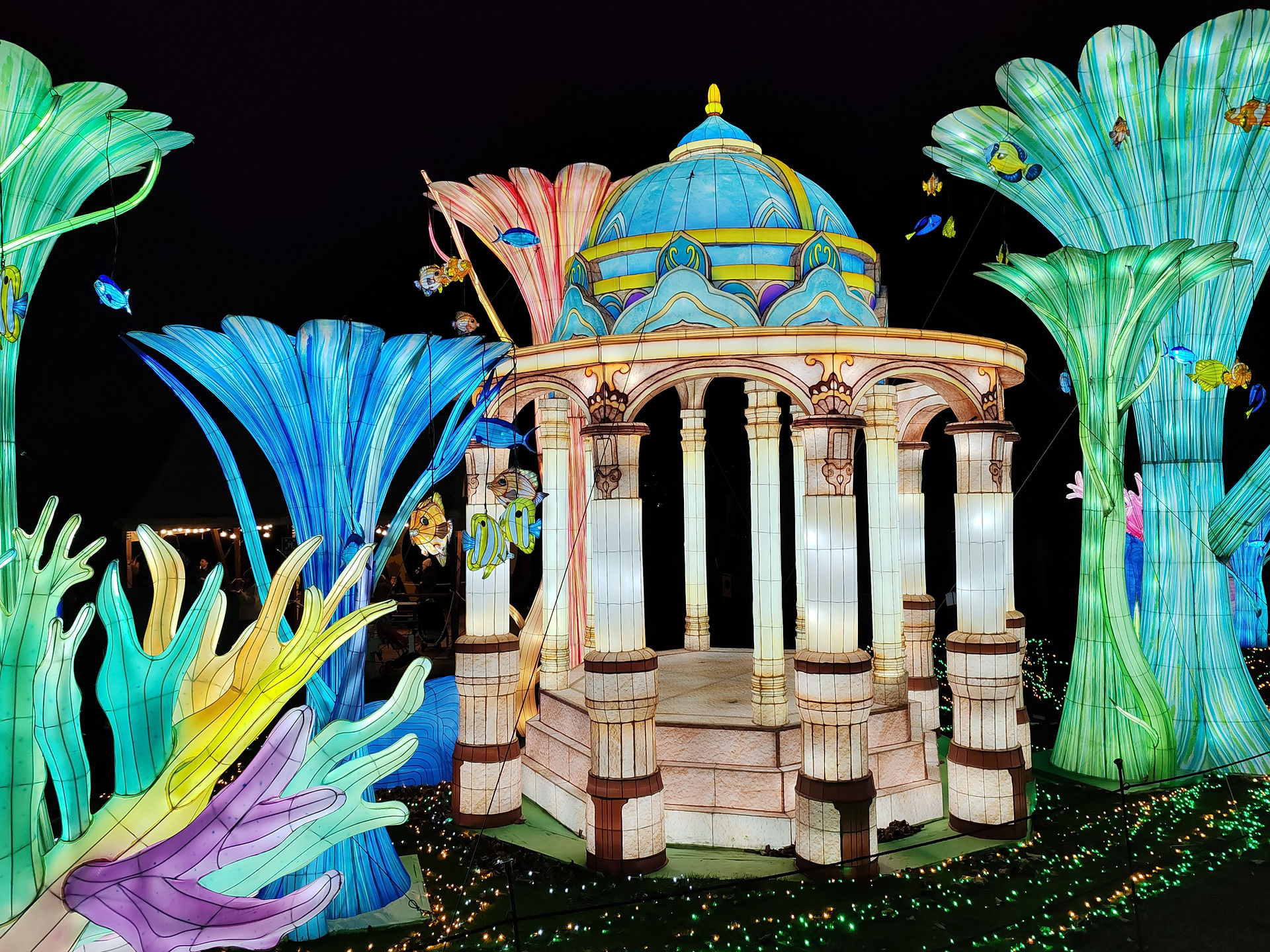 Illuminated pavilion surrounded by colourful, oversized plants and fish. Vivid blues, greens, and purples stand out against the dark night.