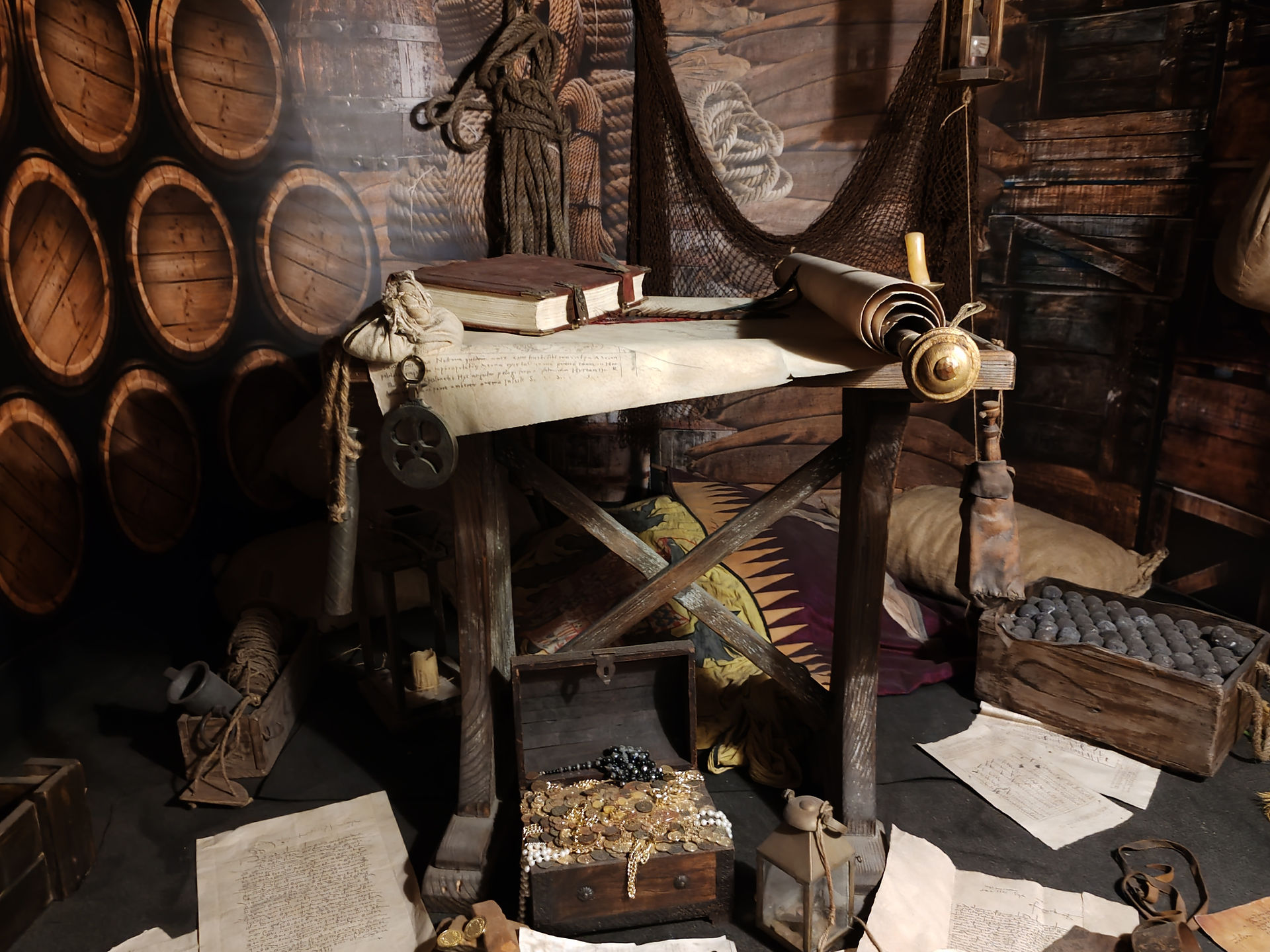 Navigation room with barrels, ropes, scrolls, and a chest of gold coins and jewels in warm, dim light.