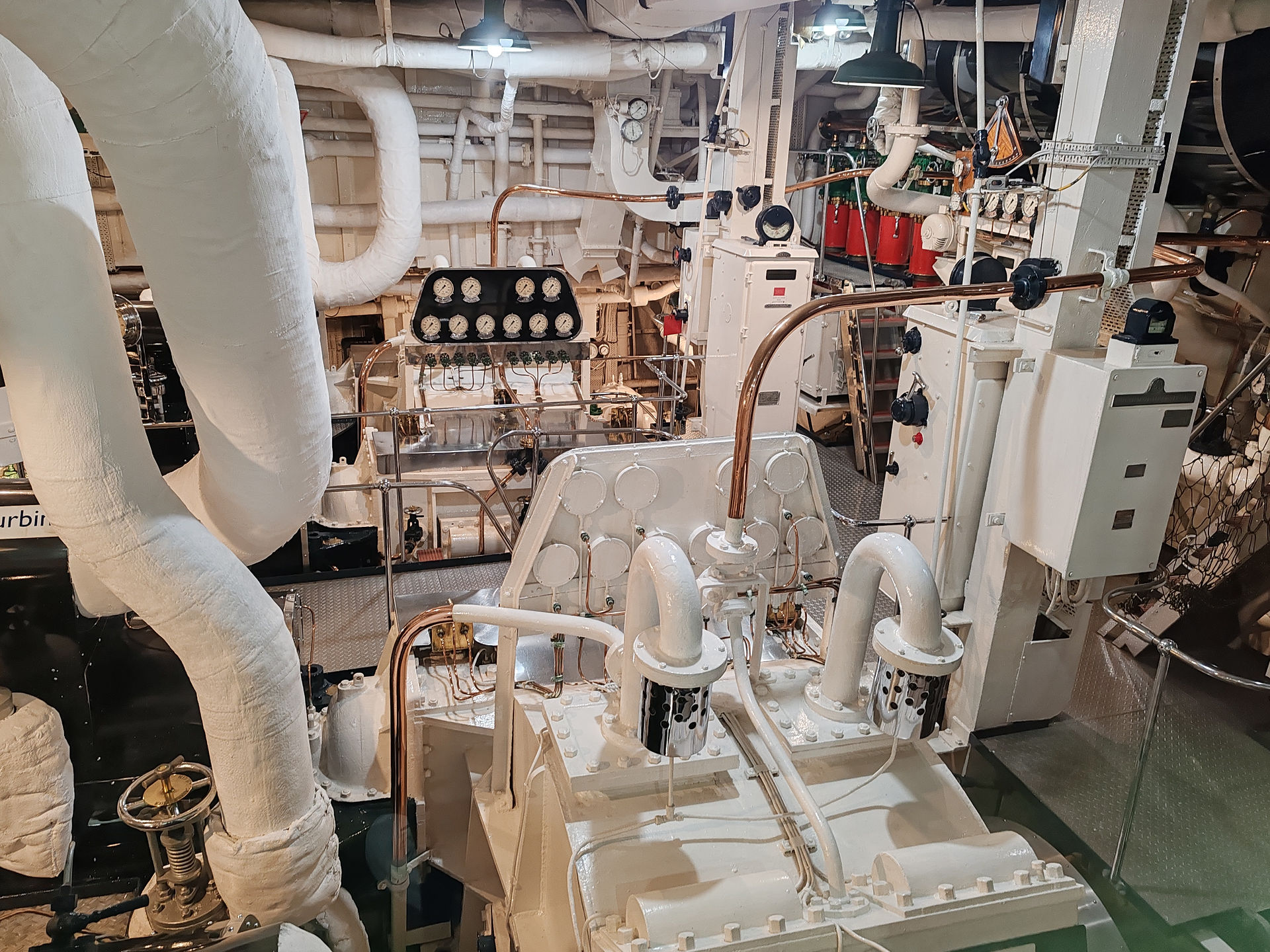 Industrial ship engine room with large white pipes, gauges, and machinery. Metal surfaces and red cylinders are visible. Bright lighting.
