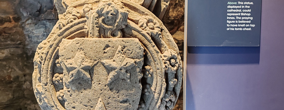 Stone carving of a vault boss with heraldry, displayed on a stand. Text panel explains historical context. Rough stone wall in background.