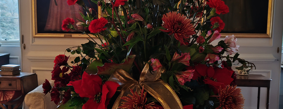 Elegant floral arrangement with red flowers and a gold ribbon sits on a table. Portraits hang on the wall in the background.