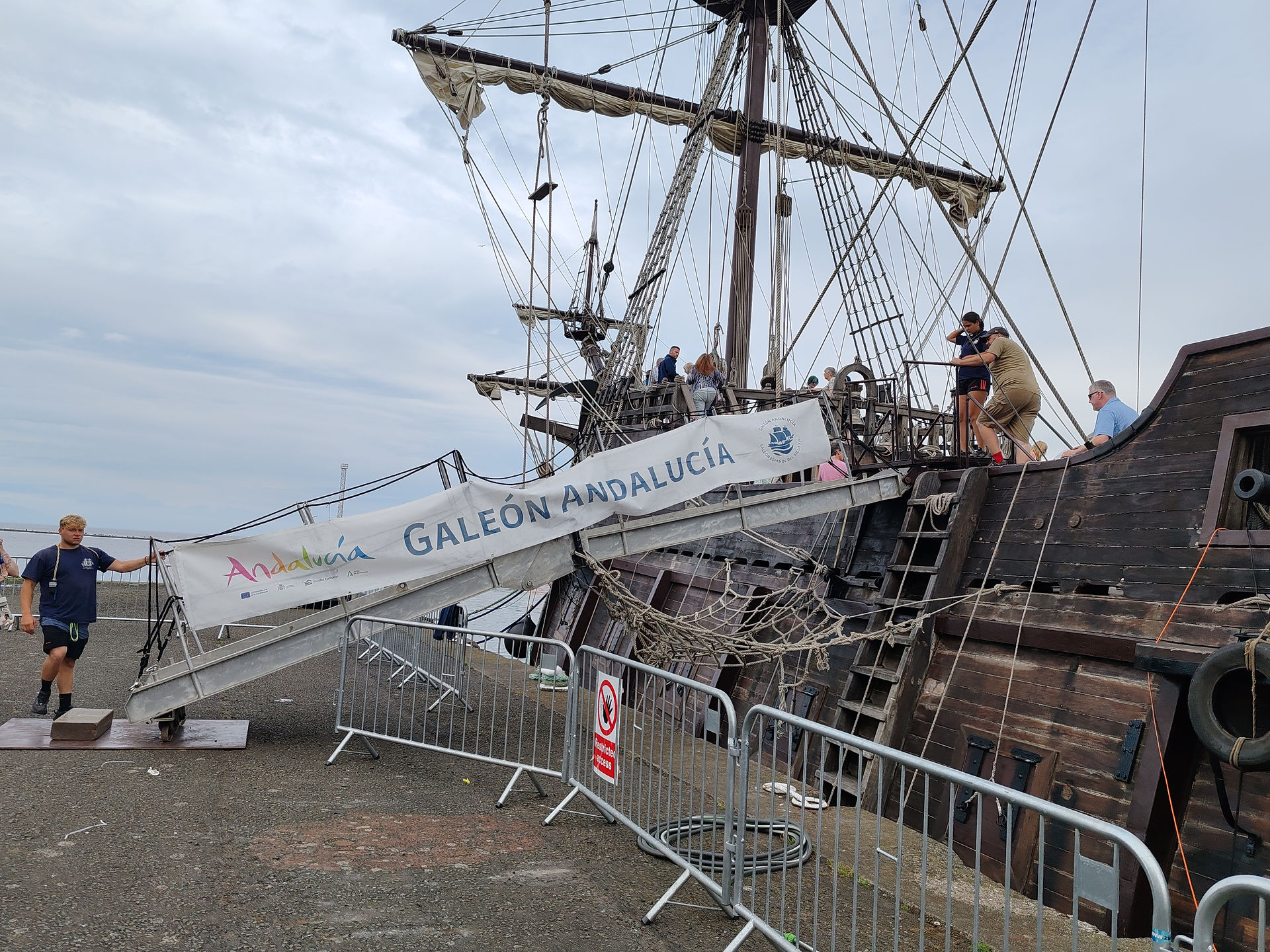 People board the replica Spanish Galleon, Galeón Andalucía, with rigging and a banner reading GALÉON ANDALUCÍA under a cloudy sky.
