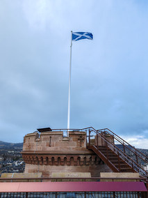 Scottish flag on a tall pole atop a stone tower at Inverness Castle with metal stairs. Overlooks a town under a cloudy sky, creating a serene mood.