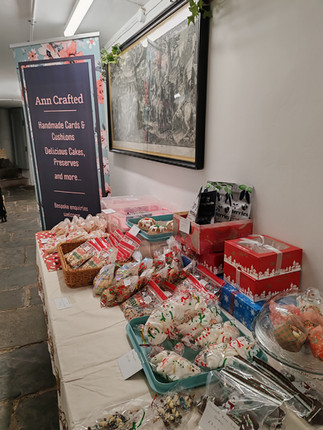 Table with festive treats and Christmas packaging displayed for sale. A sign reads "Ann Crafted: Handmade Cards & Cushions."