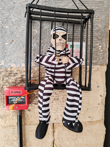 Skeleton in striped prison outfit sits in a cage, smiling. Red fire alarm nearby on stone wall. Fun, Halloween-themed décor.