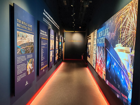 Hallway with Royal Yacht Britannia exhibits. Panels display text and images. Bright red and blue tones, fireworks image on right wall with the text, "A New Life in Leith".