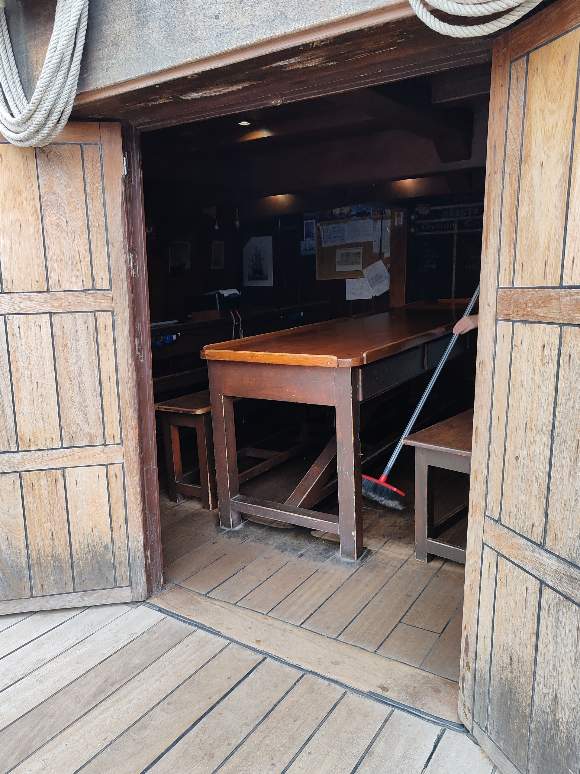 Open wooden doors reveal a dim rustic room with a long table, benches, and a hand sweeping with a broom.