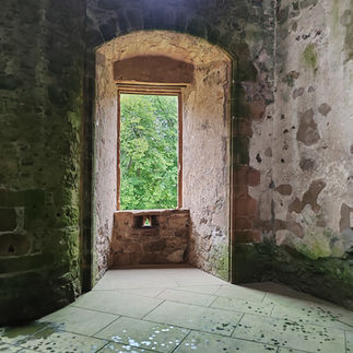 Old stone room with arched window view of green trees. Light streaming in, casting shadows on the floor. Weathered walls and floor tiles.