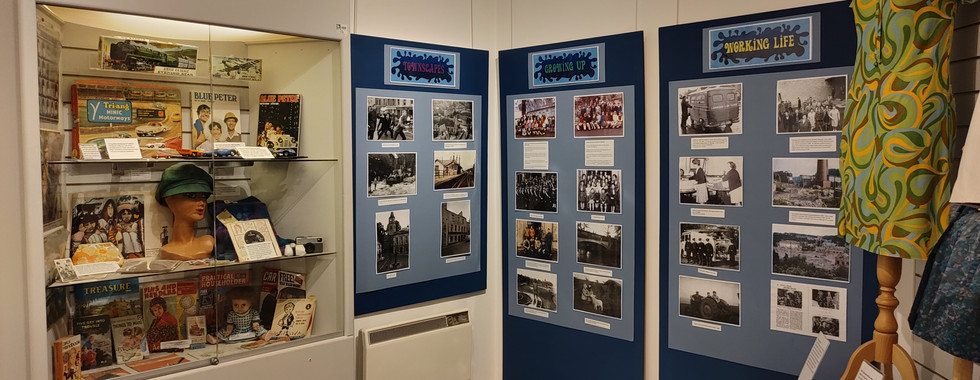 Museum exhibit with vintage items: magazines, mannequin with hat, retro photos, patterned dress on mannequin. Panels titled Growing Up, Working Life.