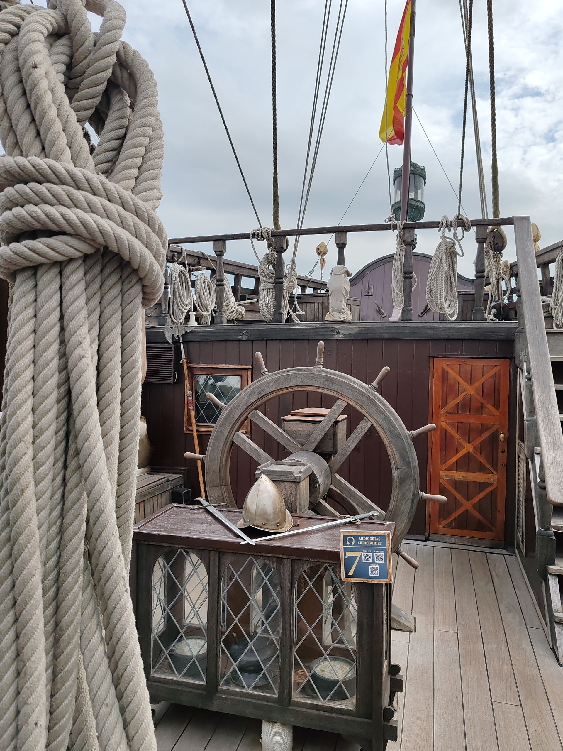 Historic ship deck with thick ropes, wooden wheel, Spanish flag, and audio guide sign under cloudy sky.