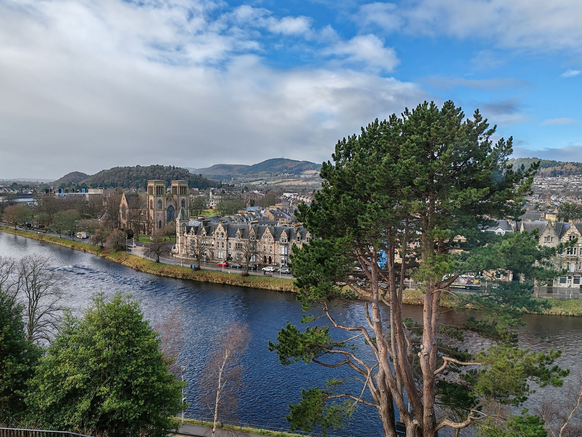 A scenic view of trees by the River Ness, with Inverness Cathedral and buildings in the background, set against hills under a partly cloudy blue sky.