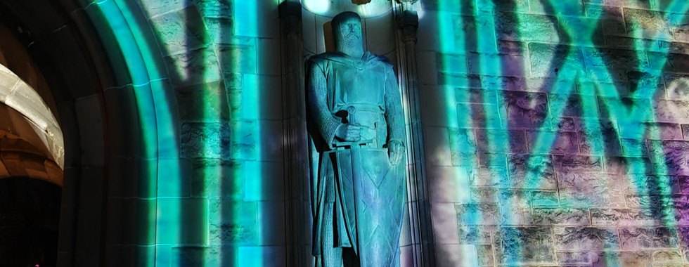 Statue illuminated by colourful green and purple patterned lights on a stone wall at night. People partially visible under archway.