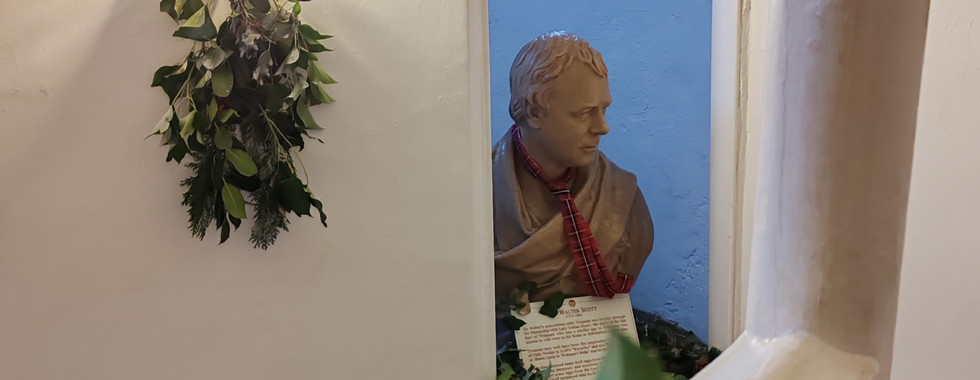 Bust of Sir Walter Scott with a red scarf, seen through a doorway. Wall sconce with greenery and berries. Text beneath bust. Warm, cosy setting.