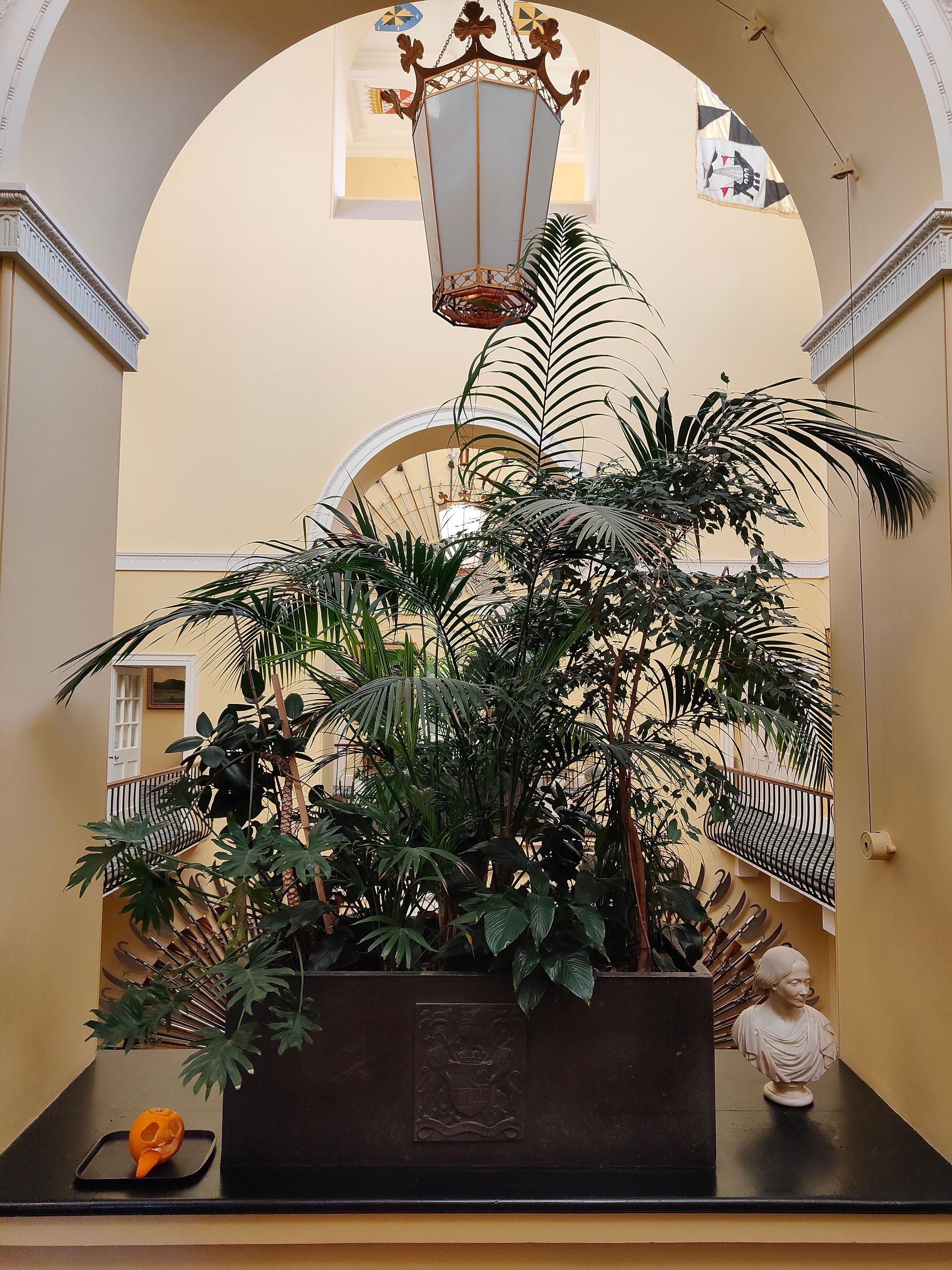 Bust near lush plants in ornate atrium. Large pendant light above, yellow walls, and decorative mouldings. Calm, elegant atmosphere.