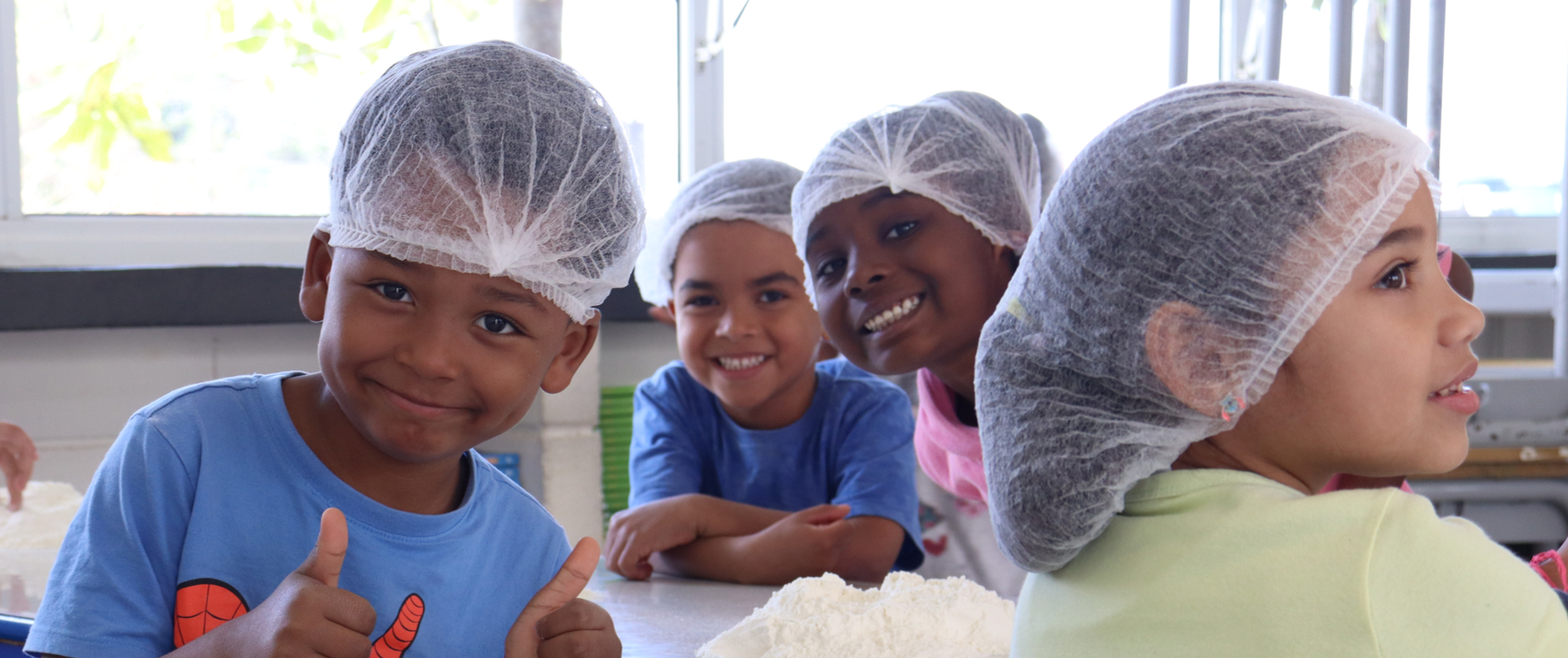 Quatro crianças sorridentes usando toucas de cozinha ao redor de uma mesa com farinha de trigo durante uma atividade prática. Em primeiro plano, um menino de camiseta azul faz sinal de joinha com as duas mãos, ilustrando como atividades lúdicas e experiências práticas nas férias promovem o aprendizado de forma leve, divertida e sem a pressão escolar.