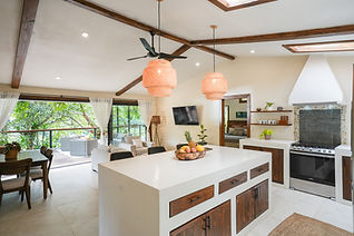 Open-plan kitchen and living area with vaulted wood-beam ceiling, center island, rattan pendant lights, and a terrace overlooking tropical gardens in Costa Rica.