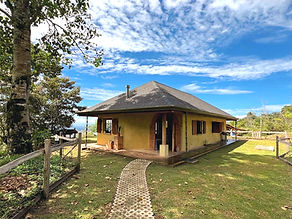 Country home in Pérez Zeledón, Costa Rica, with a covered wood-beam porch, hammock, hardwood deck, and open lawn under a blue sky.