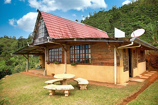 Rustic wood and stucco mountain home with red tile roof, covered porch, and stone picnic table set against forested hills in Pérez Zeledón, Costa Rica.