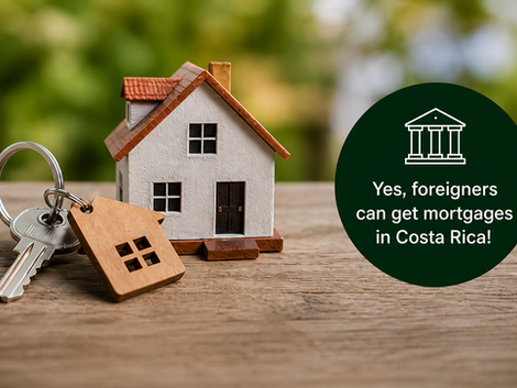 Close-up image of a small model house with a red roof sitting on a wooden surface beside a set of keys with a house-shaped keychain. A green circular badge on the right reads: “Yes, foreigners can get mortgages in Costa Rica!” against a softly blurred natural background.