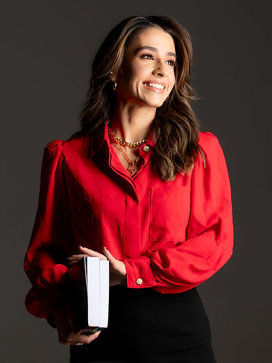 A imagem é o retrato da advogada sócia fundadora do escritório. Ela está sorrindo e segurando um livro em sua mão. Veste uma camisa vermelha e acessórios dourados.