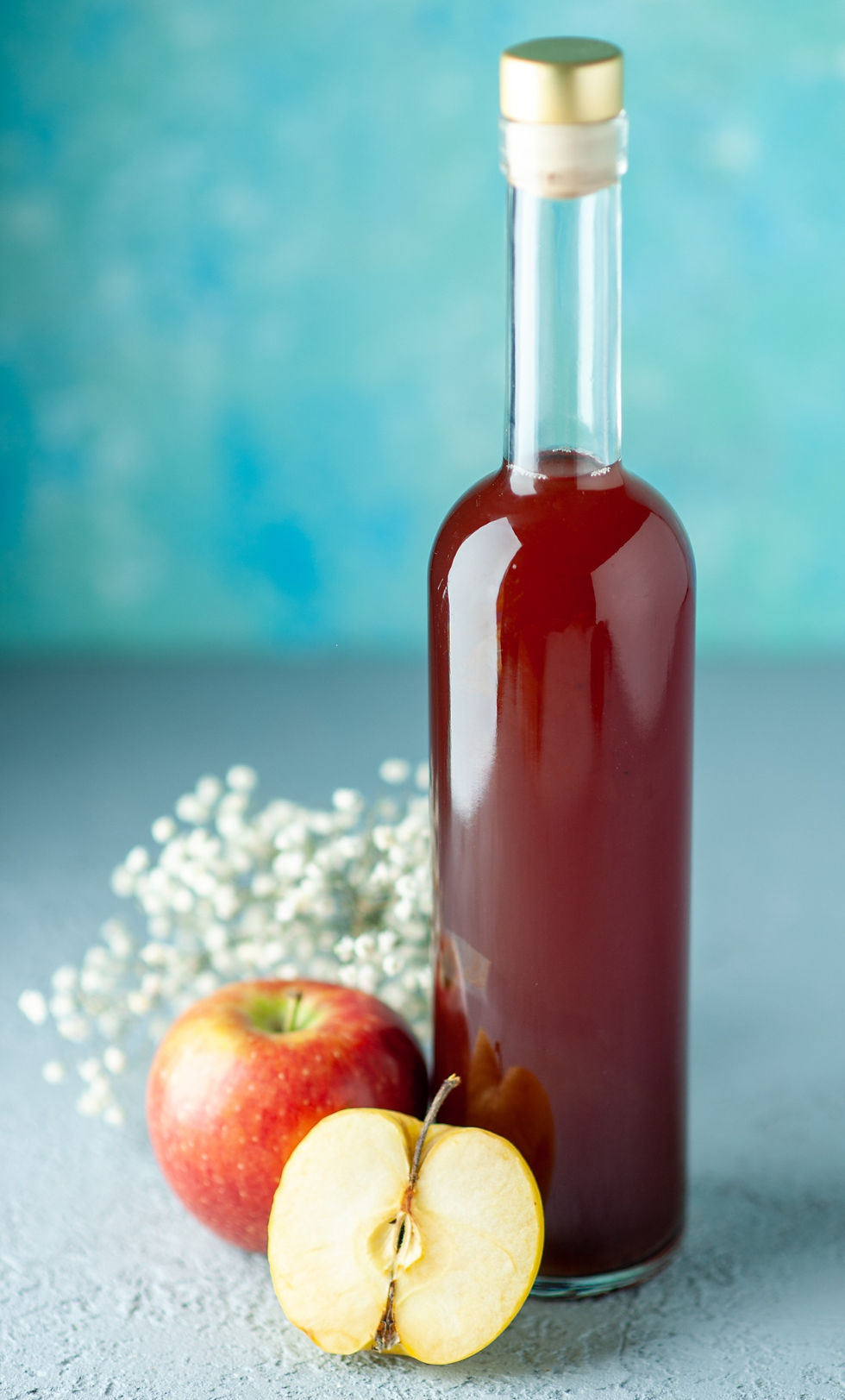 vinagre de maça caseiro, dentro de uma garraga de vidro, em frente a um fundo azul turquesa, na frente da garrafa tem uma maçã inteira e meia maçã cortada na vertical e atrás delas uma florzinha branca