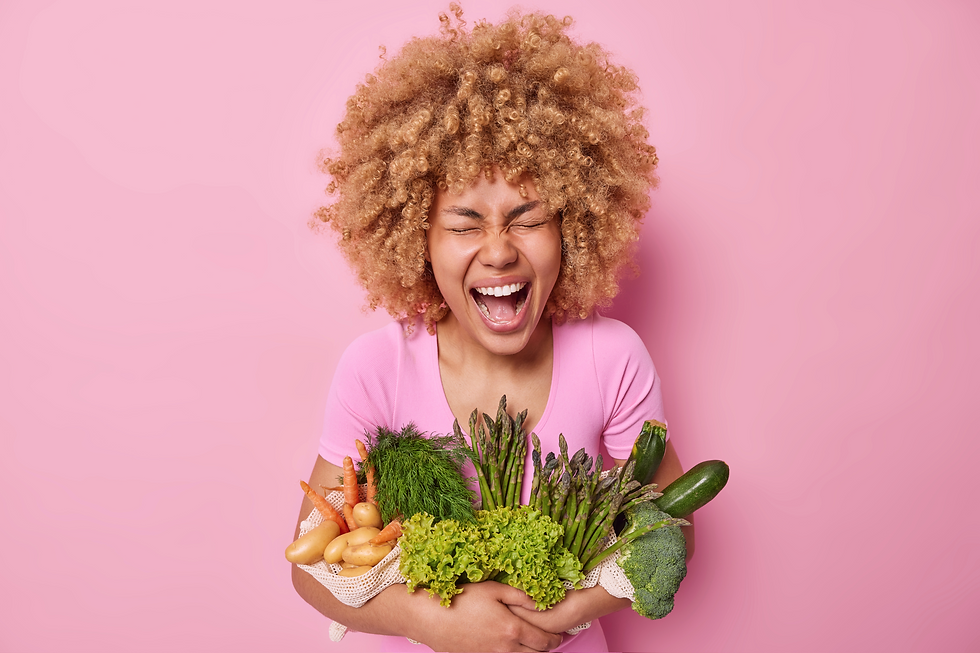 uma mulher negra de cabelos crespos, com uma expressão muito feliz, usando uma camiseta rosa, segurando uma série de legumes cruz