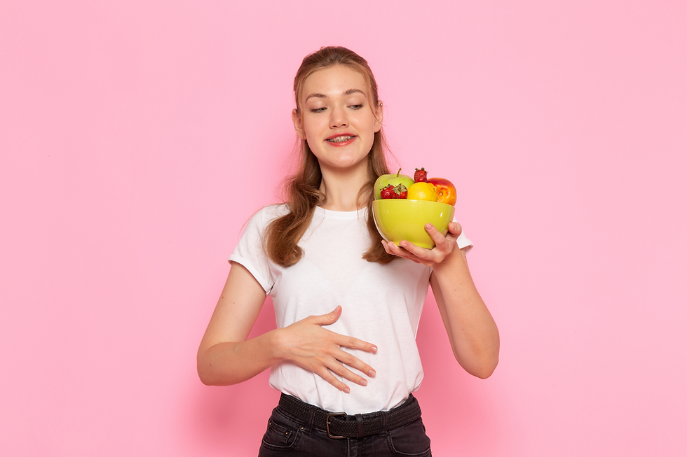 Mulher jovem com a mão em cima do ventre segurando um bowl de frutas. Ela veste calça preta e camiseta branca e em frente a uma parede rosa.