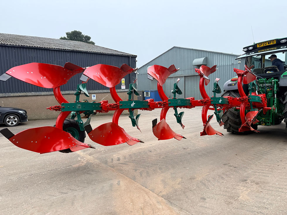 Wide landscape photo of a bright red multi-furrow plough attached to a green tractor in a farmyard