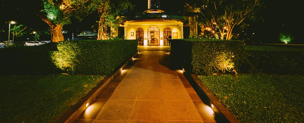 Lit garden path leads to a glowing pavilion at night, framed by hedges and trees, with a lone person by the entrance.