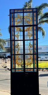 Vitral antiguo vertical con diseño vegetal dorado y marco en hierro negro, ideal para puertas o panel decorativo. Disponible en El Mundo de La Pinotea.