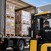 Forklift operator loading palletised POS display cartons into a freight truck at a warehouse loading dock.